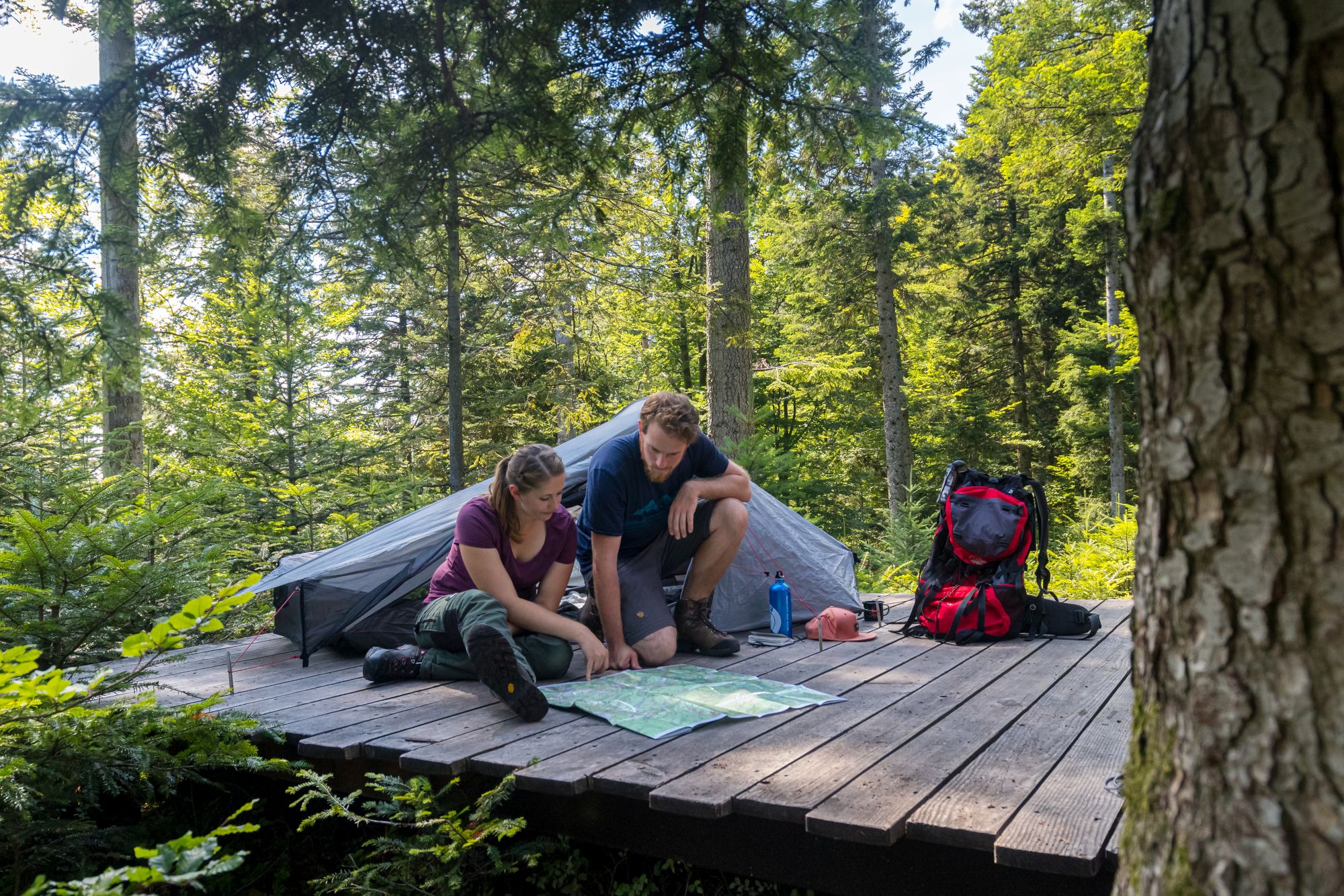 Ein Mann und eine Frau sitzen bzw. knien auf einer Holzplattform in einem Trekking-Camp im Wald und schauen gemeinsam auf eine Wanderkarte. Im Hintergrund sieht man ihr aufgebautes Zelt und einen Rucksack. (c) Sebastian Schröder-Esch Ein Mann und eine Frau sitzen bzw. knien auf einer Holzplattform in einem Trekking-Camp im Wald und schauen gemeinsam auf eine Wanderkarte. Im Hintergrund sieht man ihr aufgebautes Zelt und einen Rucksack.